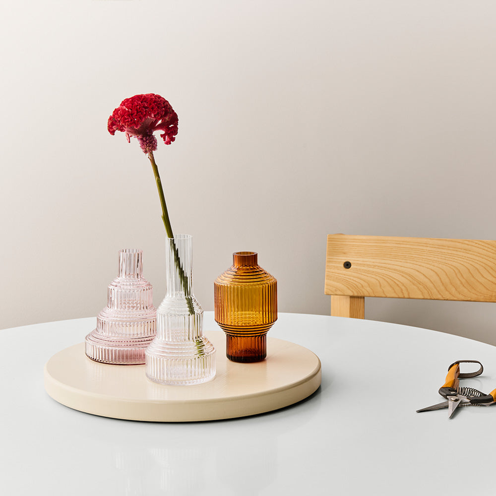 Three glass vases on a round stand with a red flower, scissor on a light surface.