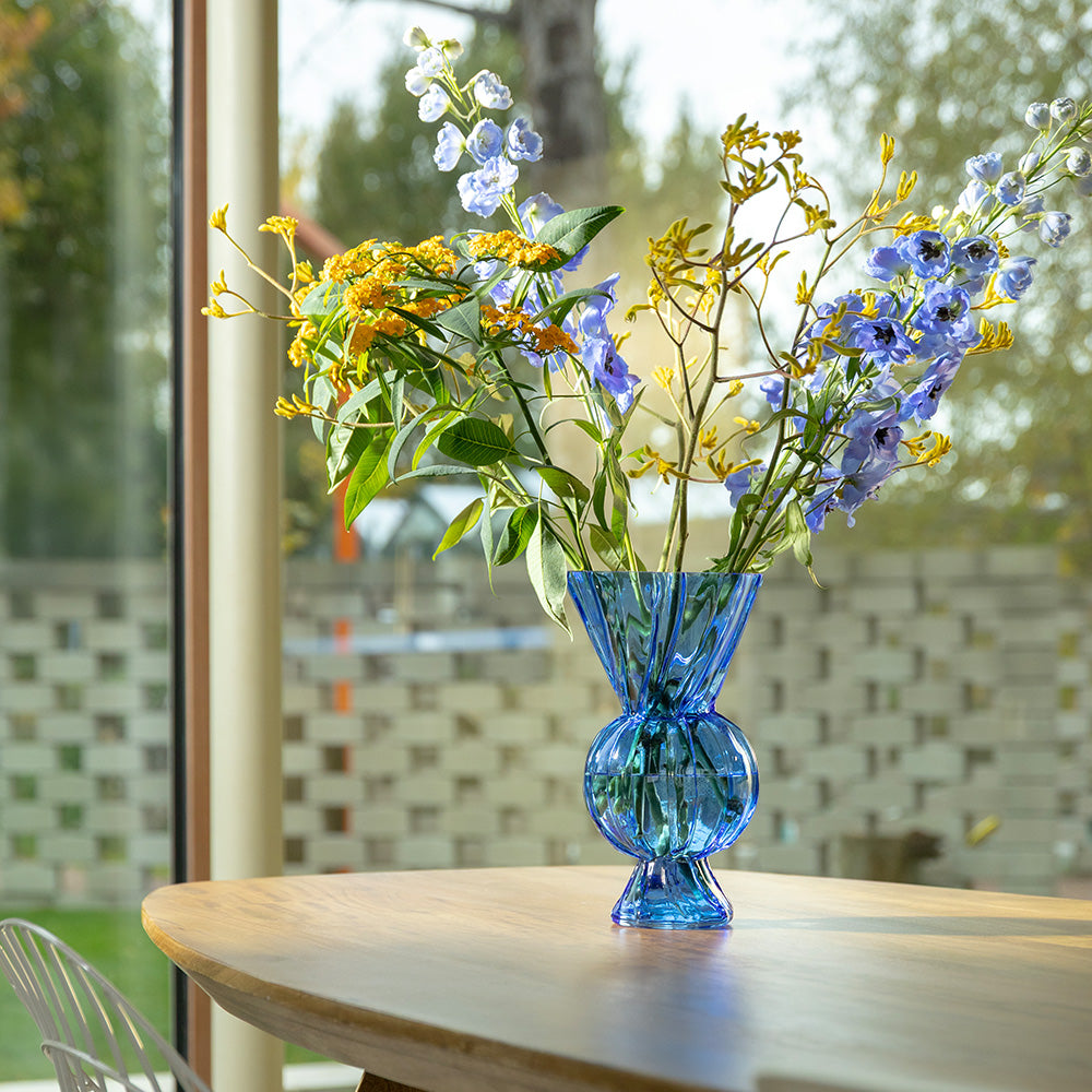 Blue vase with flowers on a table in front of a window.