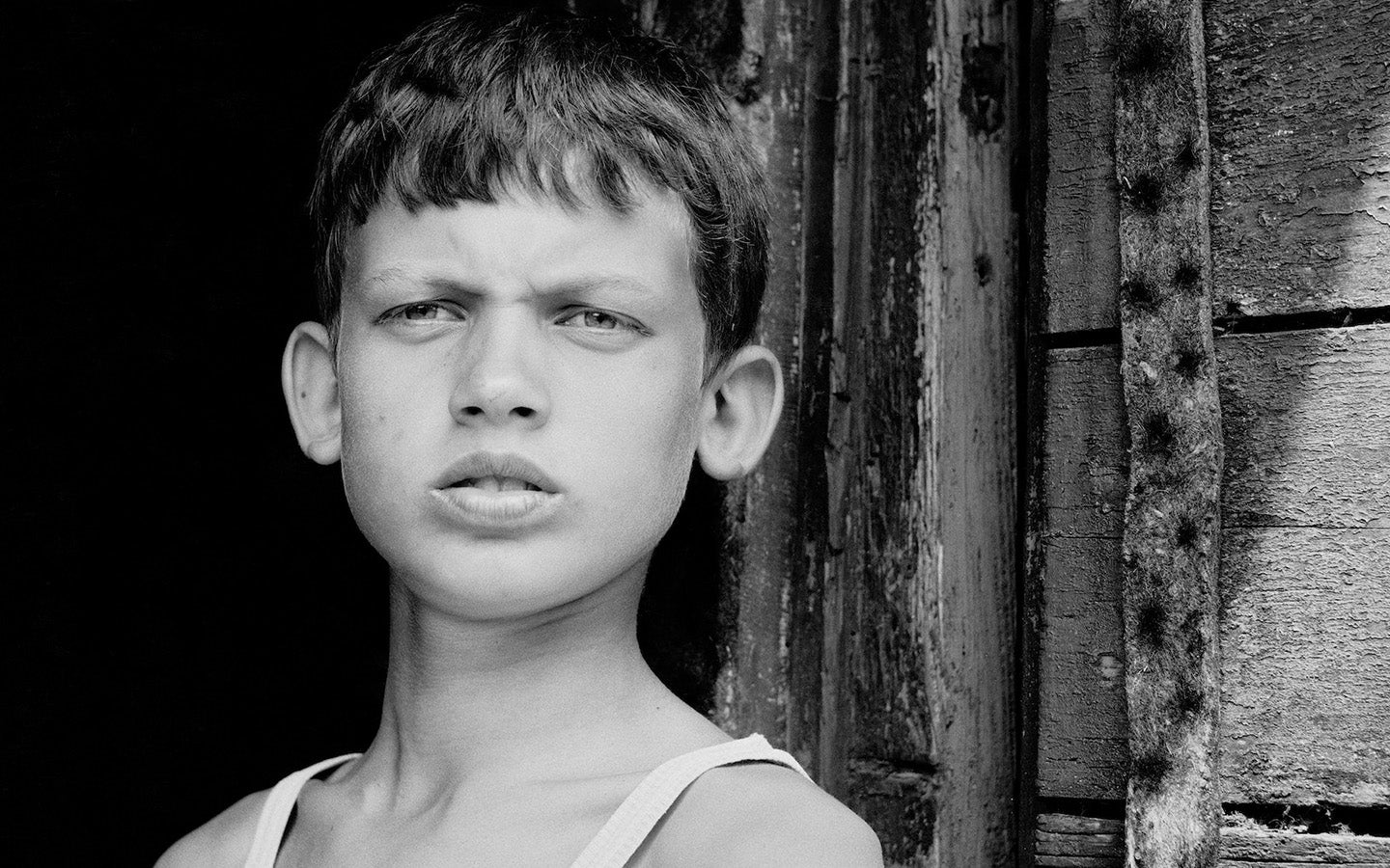Poster with a black and white motif of a young boy standing against a wooden wall by Inta Ruka.